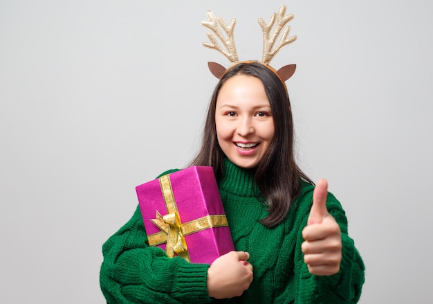 Carina giovane donna divertente vestita come un cervo di Natale esulta con un regalo su uno sfondo chiaro