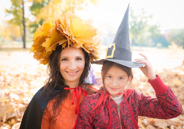 Carina giovane madre allegra con una corona di foglie di acero giallo sulla sua testa con la sua piccola figlia affascinante In un cappello da strega di carta mentre si cammina nel parco in autunno. Concetto di riunione di Halloween