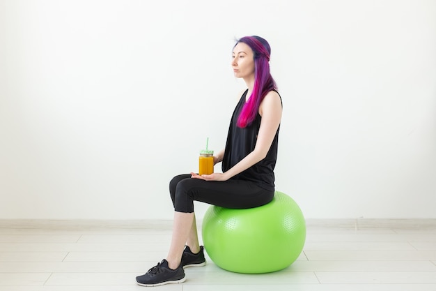Carina giovane ragazza hipster di razza mista con i capelli colorati seduto su un fitball verde e con in mano un frullato di proteine di banana su uno sfondo bianco Mangiare sano e concetto di esercizio. Copyspace