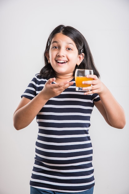 Carina piccola ragazza indiana o asiatica allegra che beve mango fresco o succo d'arancia o bevanda fredda o bevanda in un bicchiere, isolato su sfondo bianco