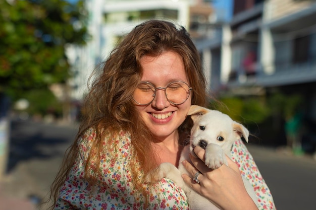 Carina ragazza allegra con cucciolo bianco tra le braccia piccolo cane cammina per strada in un giorno d'estate