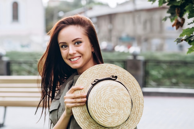 Carina ragazza bruna in un cappello di paglia all'aperto, ragazza sorridente cammina per la città in estate