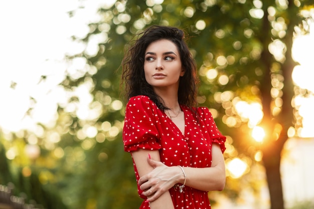 Carina ragazza di moda riccia con un bel viso in un abito rosso retrò estivo cammina nel parco al tramonto