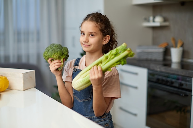 Carina ragazza ridente e riccia sta vicino al tavolo in cucina, tenendo in mano le verdure verdi.