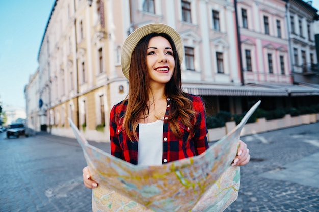 Carina ragazza turistica con capelli castani che indossa un cappello e camicia rossa, tenendo la mappa al vecchio sfondo della città europea e sorridente, in viaggio, ritratto.