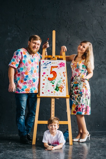 Carina, sorridente, tutta la famiglia con un pennello tra le mani disegna su un cavalletto. Il concetto di educazione, talento, una famiglia felice o genitorialità.