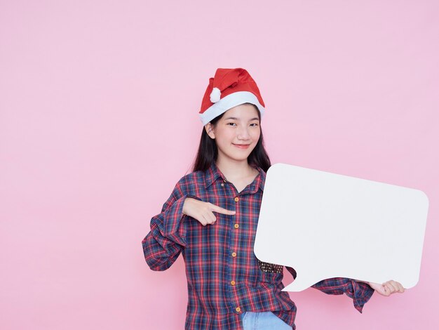 Carino adolescente gir in santa cappello puntare il dito sul poster in bianco mentre si sta in piedi su sfondo rosa.