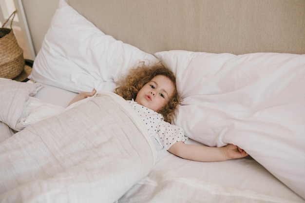 Carino affascinante bambina dai capelli ricci sdraiata a letto su un cuscino bianco.