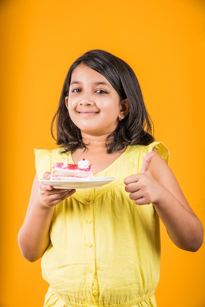 Carino bambina indiana o asiatica che mangia un pezzo di pasta o torta al gusto di fragola o cioccolato in un piatto. Isolato su sfondo colorato