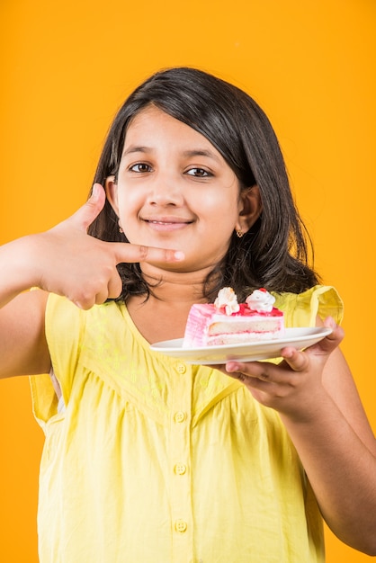 Carino bambina indiana o asiatica che mangia un pezzo di pasta o torta al gusto di fragola o cioccolato in un piatto. Isolato su sfondo colorato
