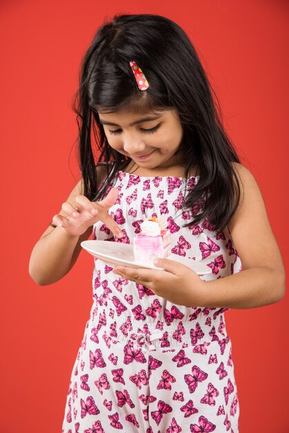 Carino bambina indiana o asiatica che mangia un pezzo di pasta o torta al gusto di fragola o cioccolato in un piatto. Isolato su sfondo colorato