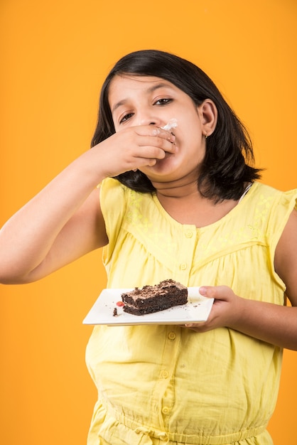 Carino bambina indiana o asiatica che mangia un pezzo di pasta o torta al gusto di fragola o cioccolato in un piatto. Isolato su sfondo colorato