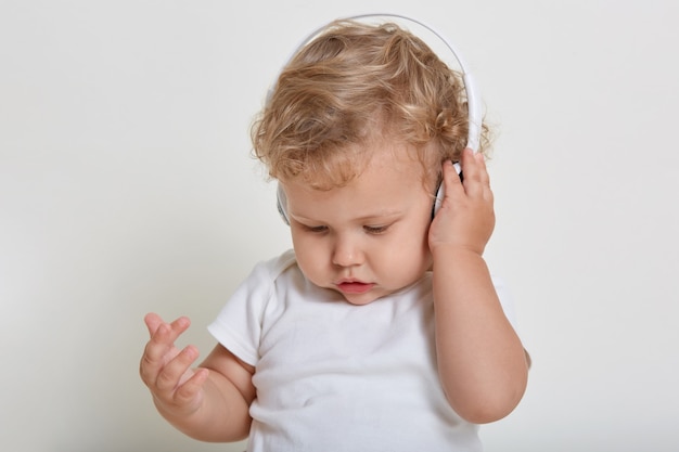 Carino bambino concentrato con capelli biondi ricci, ragazzo che indossa la maglietta, guardando lontano, toccando la cuffia con il palmo