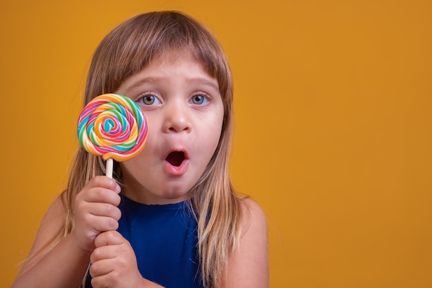 Carino bambino felice con caramelle lecca-lecca, ragazza felice che mangia grande lecca-lecca di zucchero, bambino mangia dolci. bambino sorpreso con caramelle. isolato su sfondo giallo, studio. Bella bambina con lecca-lecca