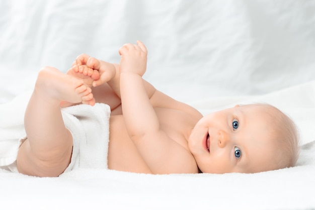 Carino bambino felice con gli occhi azzurri sdraiato sul letto bianco e tenendo le gambe Ragazzino sorridente