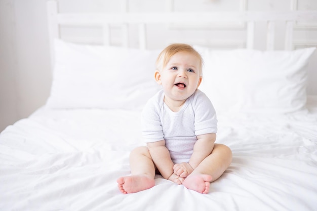 Carino bambino sorridente è seduto su un letto bianco a casa un piccolo bambino biondo si è svegliato la mattina felice infanzia e famiglia