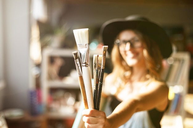 Carino caucasico artista femminile dai capelli lunghi in carino cappello nero che tiene la spazzola in uno studio contemporaneo leggero