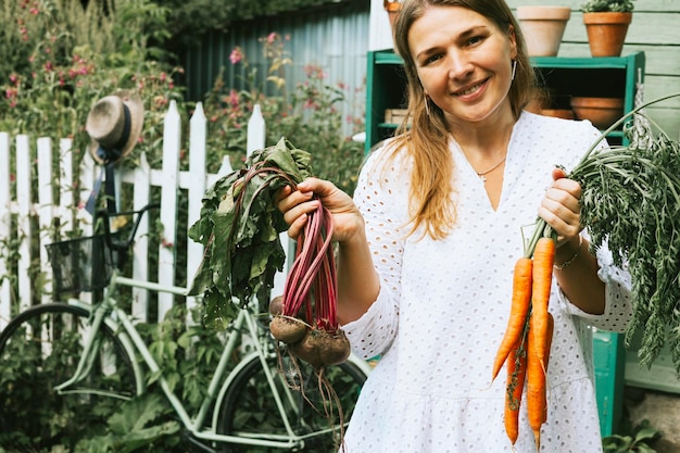 Carino contadino plus size vera donna in abito bianco casual sulla veranda della casa del villaggio di campagna con carote mature e barbabietole verdure cibo ecologico dal giardino ricco raccolto autunnale