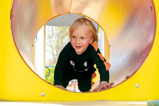 Carino divertente biondo piccolo bambino bambino bambino ragazzo che striscia arrampicata attraverso il tubo di plastica giallo nel parco giochi per bambini lo sviluppo fisico e l'asilo nido d'infanzia concetto di asilo nido