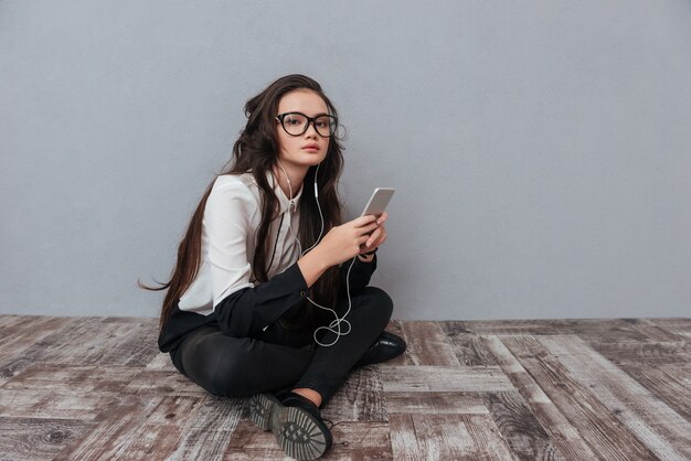 Carino donna asiatica con gli occhiali seduto sul pavimento di legno con il telefono e l'ascolto di musica.