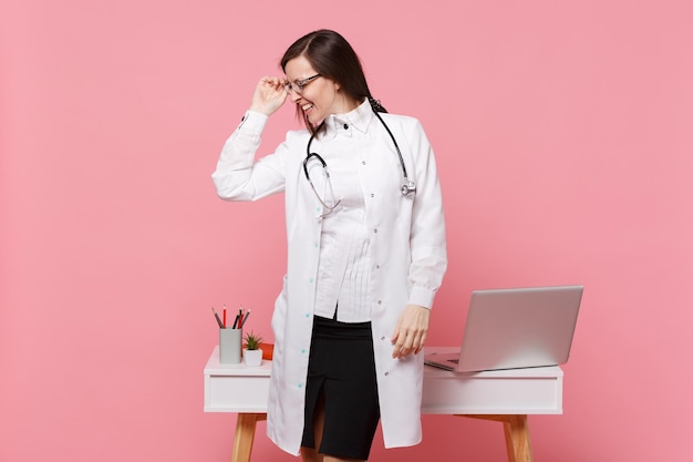 Carino dottoressa in piedi davanti alla scrivania con computer pc, documento medico in ospedale isolato su sfondo muro rosa pastello. Donna in abito medico occhiali stetoscopio. Concetto di medicina sanitaria