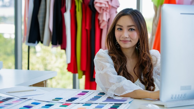 Carino ed energico designer asiatico femminile che lavora alla scrivania nella sala design tra vestiti e campioni di colore del tessuto.