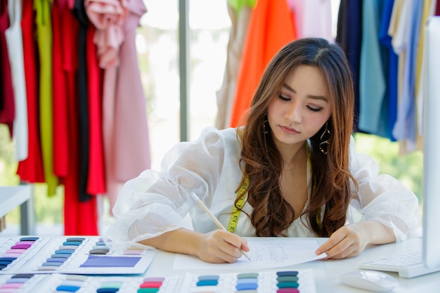 Carino ed energico designer asiatico femminile che lavora alla scrivania nella sala design tra vestiti e campioni di colore del tessuto.