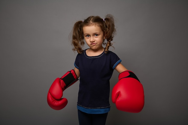 Carino europeo bambina di 4 anni che indossa guanti da boxe rossi posa su sfondo grigio muro con spazio copia per la pubblicità per il Boxing Day