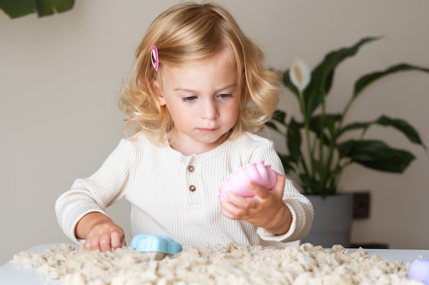 Carino felice caucasicobiondabimbo dai capelli riccibambina che gioca con la sabbia cinetica al chiusoSviluppo precoce del bambino in età prescolareconcetto di abilità sensoriali motorie