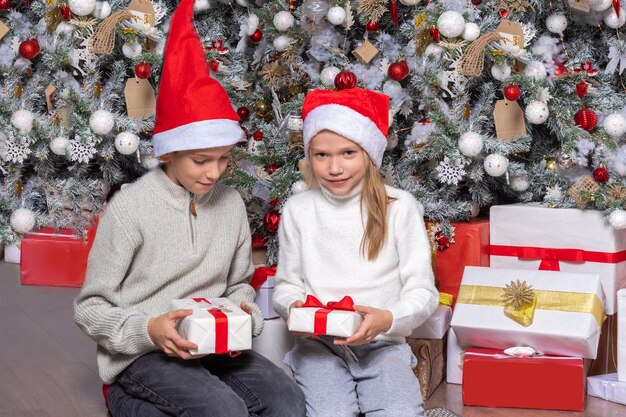 Carino felice eccitato bambini ragazzo e ragazza in cappelli di Babbo Natale si scambiano regali a sorpresa accanto all'albero di Natale a casa