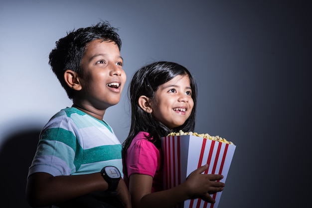 Carino felice piccolo ragazzo e ragazza indiani o fratelli che mangiano popcorn e guardano la televisione seduti su una sedia