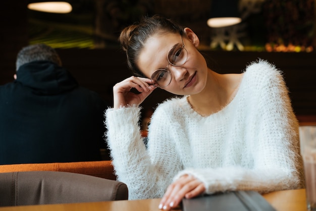 Carino giovane studentessa con gli occhiali e indossa un maglione bianco seduto in un bar dopo la scuola