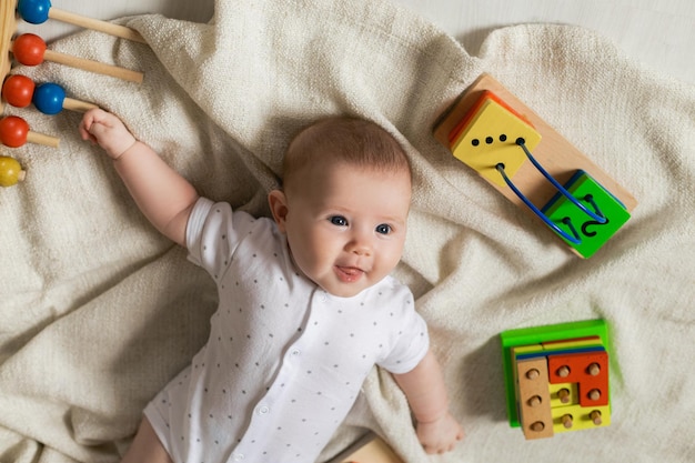 Carino neonato in abiti leggeri sta giocando con giocattoli educativi sdraiato sul pavimento su una coperta di lino grigio. vista dall'alto. prodotti per bambini. felice infanzia e maternità. spazio per il testo