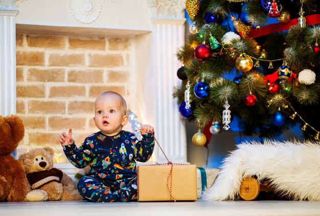 Carino piccolo bambino bambino seduto a un interno di casa sullo sfondo dell'albero di Natale con un regalo