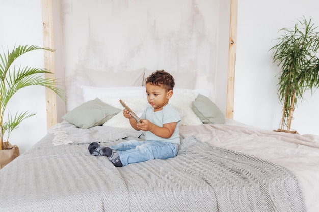 Carino piccolo ragazzo afroamericano con lo smartphone a casa sul letto bambino felice che guarda i cartoni animati sul telefono