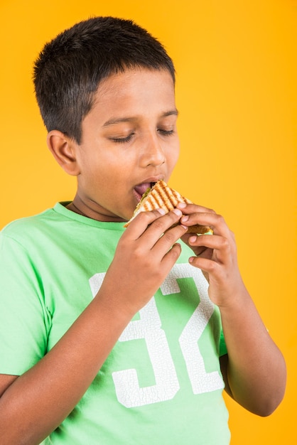 Carino piccolo ragazzo indiano o asiatico che mangia un gustoso hamburger, panino o pizza in un piatto o in una scatola. In piedi isolato su sfondo blu o giallo.