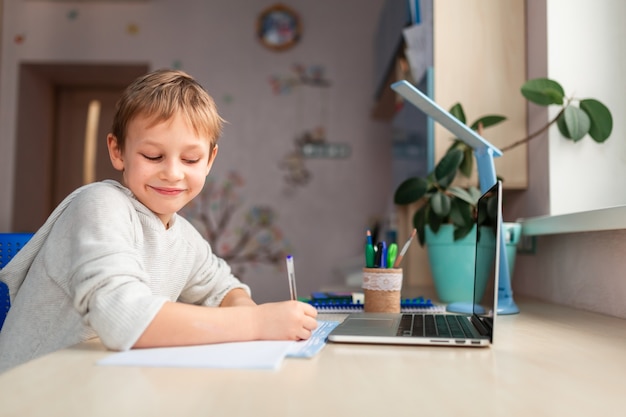 Carino piccolo scolaro felice che studia a casa facendo i compiti di scuola. Libri di formazione e taccuino sul tavolo. Formazione online a distanza