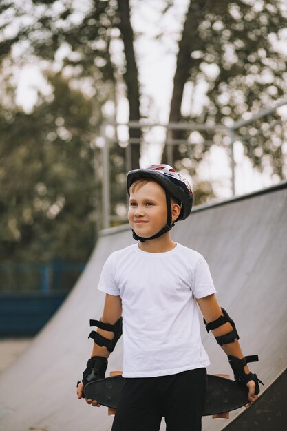 Carino ragazzo bambino in un casco in piedi in un'area speciale nello skatepark e tenendo lo skateboard Concetto di attività sportiva estiva Infanzia felice