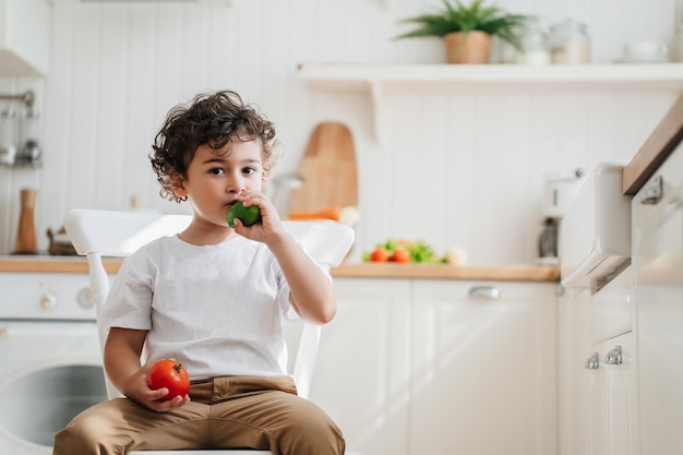 Carino ragazzo caucasico in pantaloni che mangia cetriolo tenendo il pomodoro guardando da parte Concetto di cibo sano Ricci spagnolo bel ragazzo in cucina mangia verdure Bambini in età prescolare che crescono Mangiare sano