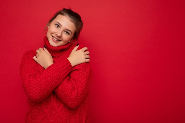 carino sorridente giovane donna che indossa un caldo maglione rosso isolato sul muro rosso