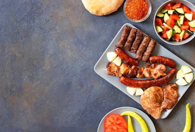 Carne alla griglia tradizionale serba e balcanica chiamata mesano meso. Barbecue dei Balcani (rostilj) servito con insalata serba, peperoncino, pane, pomodoro, cipolle e paprika in polvere. Sfondo scuro Vista dall'alto