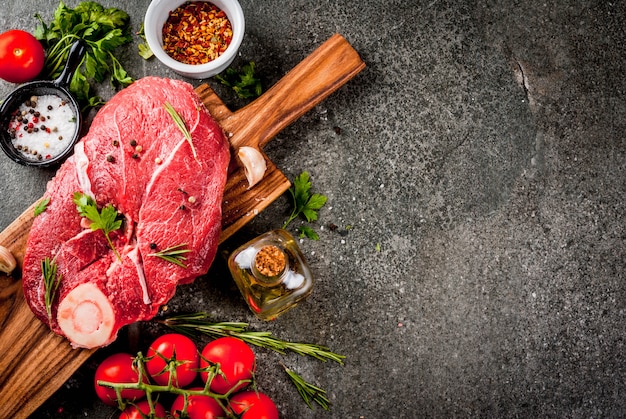 Carne cruda con ingredienti per la cena. Filetto di manzo, filetto, sul tagliere, con sale, pepe, prezzemolo, rosmarino, olio, aglio, pomodoro, funghi Tavolo in pietra nera, copia spazio vista dall'alto