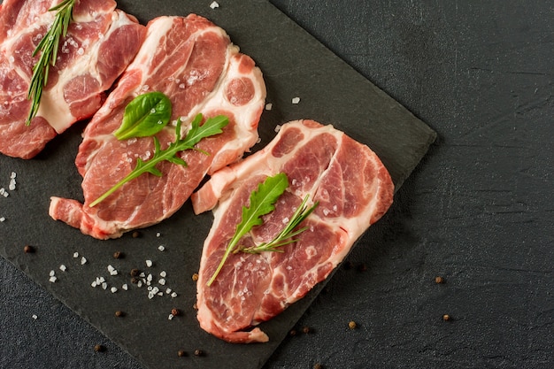 Carne di braciola di maiale cruda fresca su un'ardesia del plack con rosmarino e prezzemolo e pepe nero.