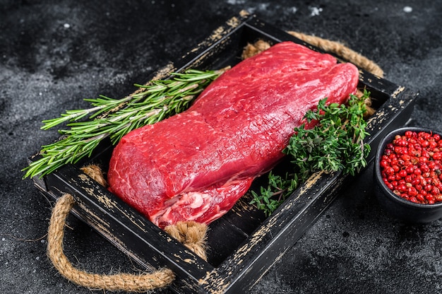 Carne di manzo intero filetto crudo su un vassoio di legno con erbe aromatiche. Sfondo nero. Vista dall'alto.