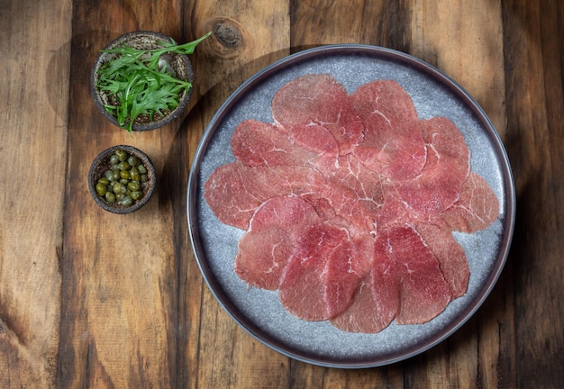 Carne di manzo marmorizzata per carpaccio con ingredienti rucola e capperi su piastra grigia in legno vista dall'alto