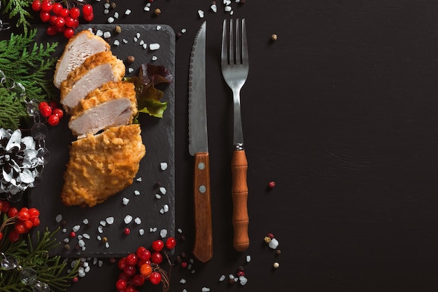 Carne di pollo bianca fritta in pastella su sfondo nero con decorazioni natalizie e posate