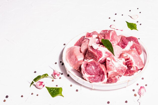 Carne di suino cruda a dadini con sommacco secco, aglio fresco e foglie di basilico. Concetto di cucina sana, luce dura alla moda, ombra scura. Sfondo bianco stucco, copia spazio