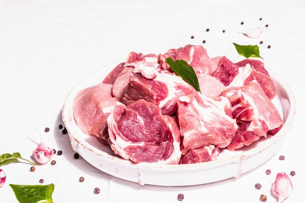 Carne di suino cruda a dadini con sommacco secco, aglio fresco e foglie di basilico. Concetto di cucina sana, luce dura alla moda, ombra scura. Sfondo bianco stucco, primo piano