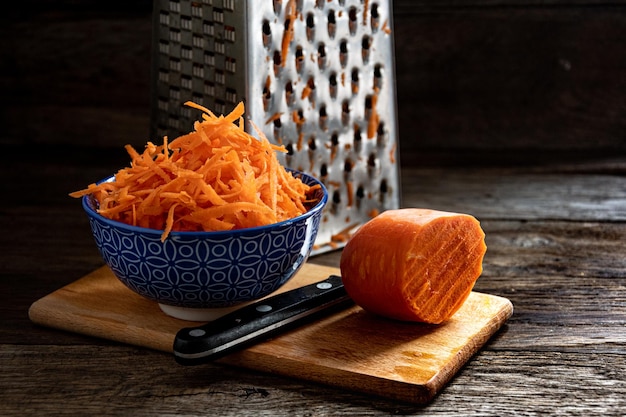 Carote grattugiate in una ciotola blu, una grattugia, un coltello e parte di una carota intera sul tavolo della cucina.