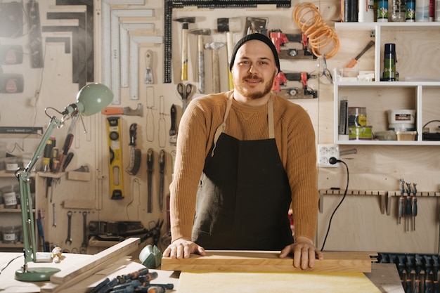 Carpentiere amichevole con un berretto da orologio in piedi dietro un tavolo in una grande officina. Numero di strumenti appesi al muro.